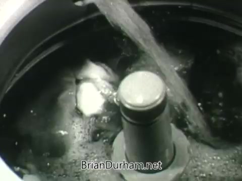 The still from the 'Public Health Service Clean Air PSA - 1960s' depicts a close-up of a spinning washing machine drum filled with water and bubbles. The focus is on the central agitator, suggesting a theme related to cleanliness and health. The visual emphasizes the importance of maintaining clean environments, likely paralleling the message of clean air awareness.