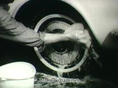 The still features a close-up of a hand washing the wheel of a car, with water and soap visibly splashing. The wheel appears to be of a mid-20th century vehicle, showcasing the cleaning process, which emphasizes care for the environment. This scene likely connects to the broader theme of public health and clean air initiatives from the 1960s.