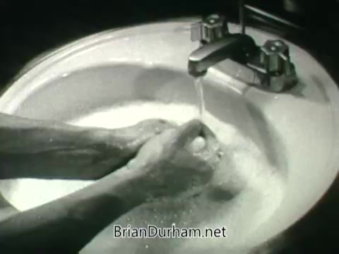 The still features a close-up of hands being washed in a sink. The water is flowing from a faucet, creating suds as the hands are scrubbed, emphasizing hygiene. The image is in black and white, typical of 1960s public service announcements promoting public health, particularly in relation to clean air and personal cleanliness.