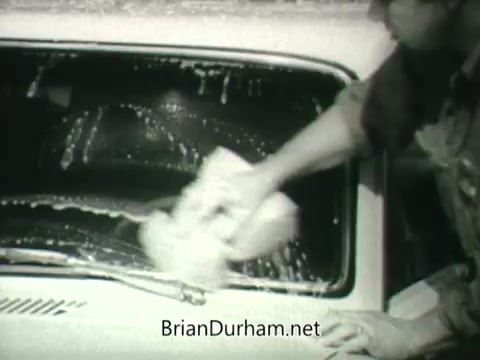 The still features a person washing a car windshield with soap and water, emphasizing cleanliness. This scene is likely part of a public service announcement promoting clean air and public health from the 1960s. The focus on cleaning suggests a connection between individual actions and broader health initiatives. The black-and-white format reflects the era’s typical style for such campaigns.