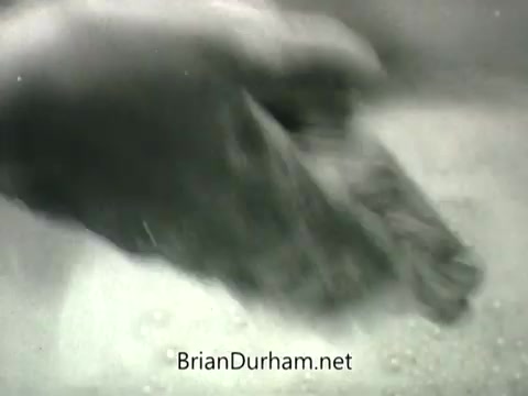 The still from the 'Public Health Service Clean Air PSA - 1960s' features a hand engaging in a washing action, submerged in soapy water. The black-and-white image emphasizes cleanliness and hygiene, reflective of the public health messaging typical of that era. It likely aims to promote awareness about air quality and health practices.