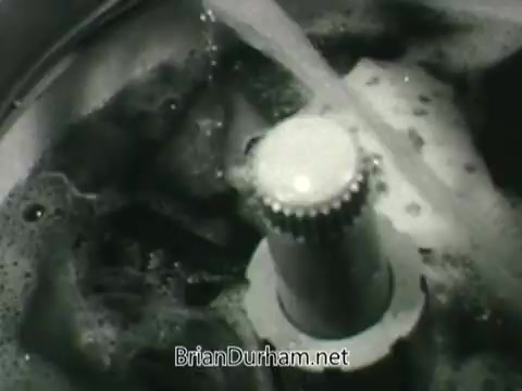 The still from the 'Public Health Service Clean Air PSA' features a close-up of a washing machine agitator in a tub filled with soapy water. Bubbles and movement suggest activity, emphasizing cleanliness and washing. This imagery likely aligns with the PSA's theme of promoting clean air and public health awareness, symbolizing the importance of a clean environment. The monochrome visual represents the era's style, typical of the 1960s.