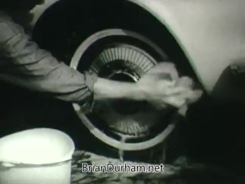 The still features a close-up of a person's hand washing a car wheel. A bucket is present nearby, indicating the cleaning process. This image is from a 1960s Public Health Service PSA promoting clean air, likely emphasizing the relationship between vehicle maintenance and air quality. The black-and-white format reflects the era's aesthetic.