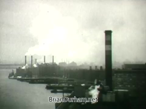The still depicts a stark industrial landscape, prominently featuring factories and smokestacks emitting thick plumes of smoke into the atmosphere. The scene likely conveys the environmental impact of industrialization, highlighting air pollution as a public health concern during the 1960s. The perspective suggests a focus on the waterways adjacent to the industrial site, emphasizing the overall grim narrative surrounding pollution and health from that era.