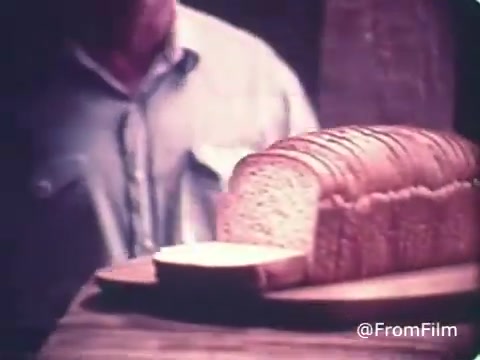 The still from the 1970s Roman Meal Bread commercial likely depicts a loaf of sliced whole-grain bread prominently displayed on a wooden board. A person, possibly a baker or homemaker, is present in the background, suggesting the theme of family and wholesome nutrition. The focus on the bread emphasizes the company's commitment to whole-grain products, aligning with its brand image of healthy eating. The visual style reflects the era, possibly featuring warm colors and a nostalgic vibe.