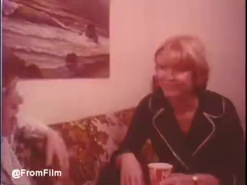 The still from the trailer of 'Alice Doesn’t Live Here Anymore' (1974) features two women engaged in conversation on a couch. One woman has blonde hair and is wearing a black outfit with a white collar, holding a cup. The setting appears casual and domestic, with a colorful patterned couch and a painting of a beach in the background. The mood seems warm and intimate, capturing a moment of connection between the characters. The trailer's music includes 'All the Young Dudes' by Mott The Hoople, adding to the film's 1970s vibe.