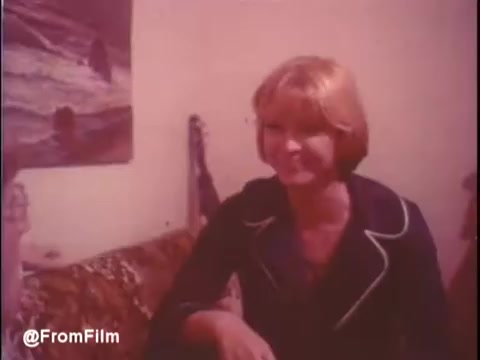 The still from the trailer of 'Alice Doesn't Live Here Anymore' features a young woman with short, blonde hair, smiling as she sits on a patterned couch. The background suggests a cozy living space, with a painting and possibly personal items visible. The atmosphere appears warm and inviting, reflecting the character's optimistic demeanor. The trailer is underscored by music from Mott The Hoople, adding to the nostalgic vibe of the film.