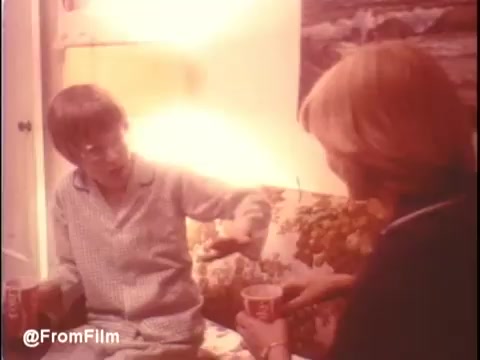 The still from 'Alice Doesn't Live Here Anymore' shows two women seated in a cozy living room, likely engaged in a conversation. One woman, dressed in pajamas, is gesturing with her hand, while the other holds a cup, appearing attentive. The warm lighting and floral-patterned couch create an intimate atmosphere, reflecting the film's themes of personal struggles and female relationships. The trailer features music by Mott The Hoople.