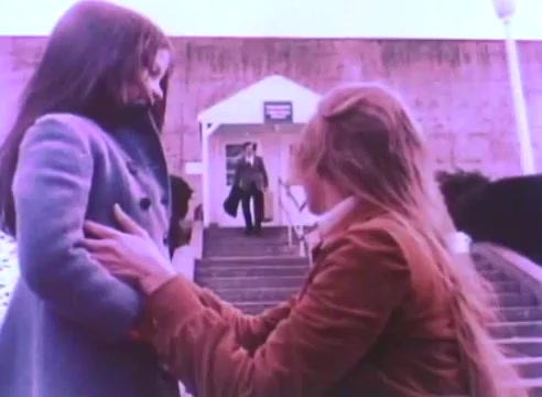 The still from the 'Hire An Ex-Offender' PSA depicts a woman kneeling beside a young girl, holding her gently while looking up. In the background, a man in a suit is seen ascending the steps of a building, suggesting a reunion or significant moment. The scene conveys a sense of emotional connection and hope, emphasizing themes of redemption and the importance of reintegrating ex-offenders into society. The color palette and film quality reflect the style of 16mm prints, contributing to a vintage aesthetic.