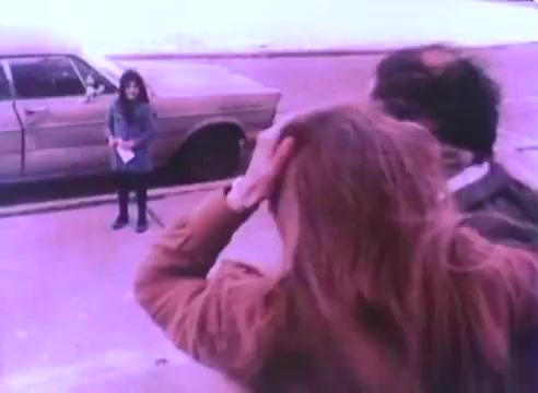 The still from the 'Hire An Ex-Offender PSA' features a scene set on a street with a parked car in the background. In the foreground, two people, a man and a woman, appear to be discussing something while a woman in a blue coat stands nearby, holding papers. The overall tone seems to convey a moment of interaction or tension, reflecting themes of employment and social perception regarding ex-offenders. The color palette and graininess suggest it is from a vintage 16mm film.