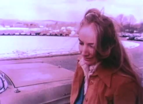 The still captures a woman with long hair, looking pensive or sad. She is wearing a brown jacket and is positioned near a car by a body of water. The color palette has a muted, vintage tone, characteristic of a 16mm print. The background features a snowy landscape and parked cars, suggesting a cold, possibly desolate setting. The mood appears reflective, aligning with the theme of the PSA focused on hiring ex-offenders.