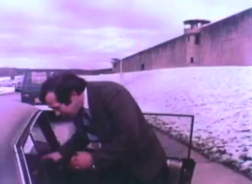 The still depicts a man in a suit leaning into a car, likely preparing to get inside. In the background, a prison wall is visible, along with a guard tower, suggesting a setting related to rehabilitation or reintegration. The overall tone is somber, reflecting the serious theme of the PSA advocating for hiring ex-offenders. The image has a dated aesthetic typical of 16mm film.