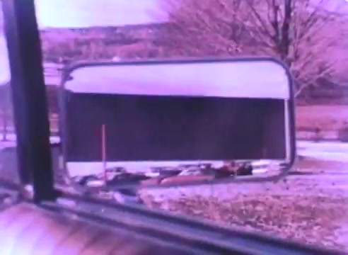 The still from the 'Hire An Ex-Offender PSA' features a reflection in a side mirror, showcasing a blurred view of a parking lot with cars. The scene is bathed in a pinkish hue, indicative of the vintage 16mm film quality. A tree can be seen in the background, adding to the outdoorsy context. The focus appears to be on the perspective of a vehicle, hinting at themes of movement and observation related to the PSA's message.