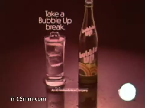 The still from the 'Bubble Up' soda commercial features a glass filled with ice and a refreshing drink, alongside a bottle of Bubble Up prominently displayed. The background is likely a warm, inviting color. Text on the image encourages viewers to 'Take a Bubble Up break,' emphasizing the brand's playful and refreshing qualities. The overall presentation conveys a sense of fun and thirst-quenching enjoyment, aligning with the upbeat voice-over promoting the beverage.