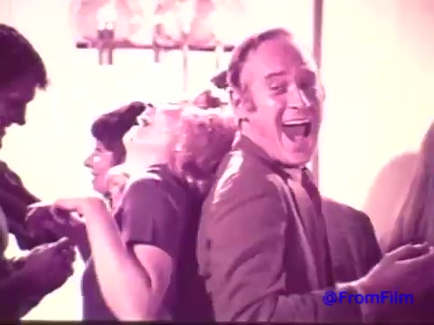 The still from the 1970s Nabisco Korker Corn Twists commercial captures a lively scene with people enjoying themselves at a social gathering. The faded print, transitioning from red to black and white, enhances the nostalgic quality of the moment. Central to the image is a man with a large, joyful grin, suggesting a fun atmosphere. Nearby, a woman leans back in laughter. The overall vibe reflects the playful and carefree spirit typical of snack advertisements from that era.