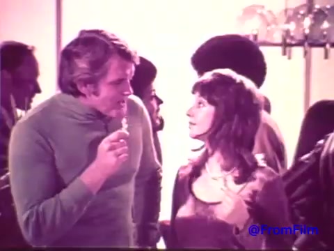 The still from the Nabisco Korker Corn Twists advertisement features a man and woman engaged in conversation at a social gathering. The man, dressed in a light green turtleneck, holds a Korker snack and appears to be playfully interacting with the woman, who wears a brown outfit. The background is filled with other figures, and the scene is characterized by a faded look due to the 16mm print's age, shifting from red tones to black and white. The overall ambiance reflects a casual, vintage 1970s party atmosphere.