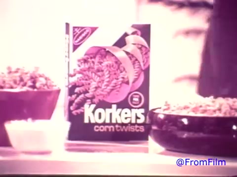 The still features a product display of Nabisco Korkers Corn Twists from the 1970s. The box is prominently shown, with its distinct design, featuring a purple color scheme now faded to a softer tone. Accompanying the box are two bowls filled with the snack, suggesting its appealing crunchiness. The image overall reflects a vintage aesthetic characteristic of that era, despite the color fade from red to black and white. The nostalgic vibe captures the essence of a short-lived snack product.