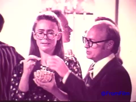 The still features a woman with large glasses holding a bowl of Nabisco Korker Corn Twists, smiling as she interacts with a man beside her, who is also wearing glasses. The background is slightly blurred, populated by other party-goers. The faded colors give the image a nostalgic quality, reminiscent of 1970s advertising. The scene captures the light-hearted social atmosphere surrounding the short-lived snack product.