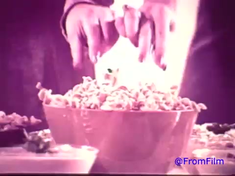 The still from the 'Nabisco Korker Corn Twists - 1970's' features a close-up of hands reaching towards a large bowl filled with twisted corn snacks. The image, originally in color, has faded to black and white, creating a nostalgic feel. Surrounding the bowl, additional snack items are visible, likely part of a larger spread. The atmosphere suggests a casual, inviting setting, possibly for a gathering or party.