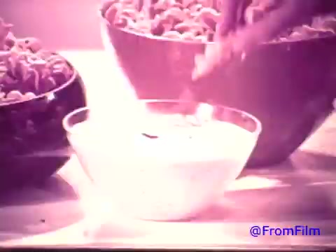 The still from the 16mm print of the 'Nabisco Korker Corn Twists' advertisement features a hand dipping a snack into a bowl of dip. There are two large bowls of Korker Corn Twists in the background. The print has faded from red to a monochromatic black and white. The scene captures a moment of enjoyment typical of snack time in the early 1970s, highlighting the product’s appeal as a fun and shareable snack.