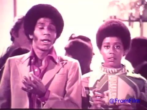 The still features two characters, a man and a boy, likely in a social setting. The man, dressed in a flamboyant outfit typical of the 1970s, appears animated while discussing something. The boy looks on, holding a snack, with a surprised or curious expression. The background suggests a casual gathering, with blurred figures indicating more people present. The print's faded quality, transitioning from red to black and white, adds a nostalgic feel, evoking the era of the short-lived Nabisco Korker Corn Twists.