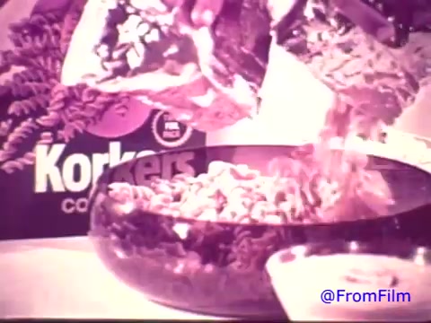The still features a close-up shot of two bags of Nabisco Korkers being poured into a clear bowl. The iconic Korkers packaging is visible in the background, showcasing its distinctive logo. The image has a faded quality, shifting from the original red hues to a monochromatic tone, characteristic of older 16mm prints. The scene captures the playful essence of 1970s snack culture.