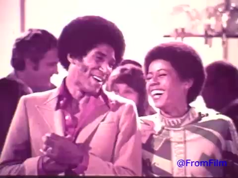 The still from the 1970's Nabisco Korker Corn Twists advertisement features two individuals, a man and a woman, sharing a joyful moment. Both are smiling and engaged in conversation, dressed in period-appropriate attire, reflecting the fashion of the era. The image, derived from a faded 16mm print, showcases a transition from red tones to black and white, giving it a nostalgic feel. The background hints at a lively social gathering, reinforcing the fun and carefree spirit of the snack.