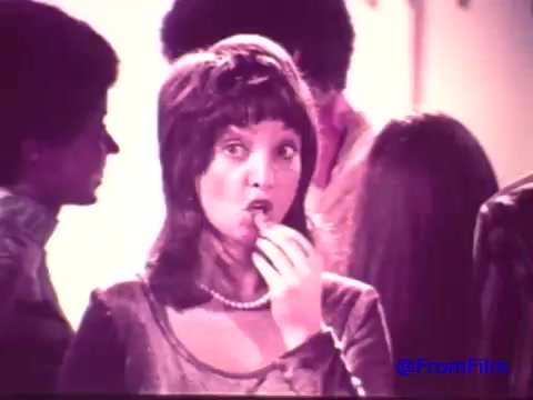 The still features a woman with a surprised expression, enjoying a Nabisco Korker Corn Twist. The faded colors suggest a nostalgic look typical of early 1970s advertising, with prominent elements like her pearl necklace and styled hair reflecting the fashion of the time. In the background, other figures are blurred, contributing to a party atmosphere. The overall tone is playful and inviting, capturing the essence of a short-lived snack chip from that era.