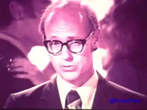 The still features a man in a suit with large, thick glasses, delivering a message directly to the camera. His expression appears earnest and slightly serious, contrasted by a blurred background where people seem to be socializing, possibly at a party or event. The image has a faded quality due to the loss of color, resulting in shades of black and white with a slight tint reminiscent of the 1970s aesthetic. The caption and branding for 'Nabisco Korker Corn Twists' suggest a nostalgic promotion for this short-lived snack.