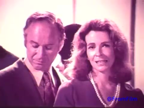 The still from the 1970s Nabisco Korker Corn Twists advertisement depicts two individuals in a promotional context. The scene, which has transitioned from its original color to a faded black and white, features a man looking slightly away while a woman speaks directly to the camera. She appears to be engaging the audience with a friendly demeanor, likely discussing the product. The overall aesthetic reflects the style and tone of advertising during that time, emphasizing personal connection.