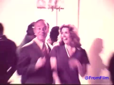 The 16mm still from the 1970s Nabisco Korker Corn Twists advertisement features a man and a woman dancing joyfully against a blurred background of partygoers. The colors have faded, giving the image a monochromatic and nostalgic quality. The man, dressed in a suit, smiles broadly alongside a woman with flowing hair, both seemingly enjoying the moment. The atmosphere suggests a lively gathering, likely promoting the fun associated with the snack.