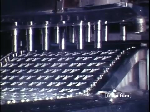 The still from 'Making Frozen Novelties - 1967 Kodachrome' features a close-up view of an industrial machine involved in the production of ice cream novelties. The scene captures a metal surface with a grid-like pattern, likely part of a molding or forming process. The shiny, metallic texture suggests the machinery's precision and the efficient production methods of the era. With the footage originating from DCA Food Industries, it highlights the technological advancements in ice cream manufacturing during the late 1960s.