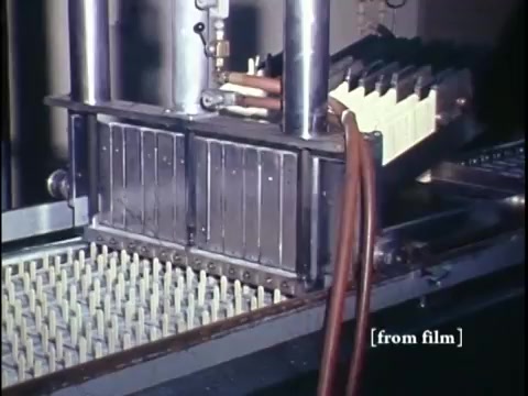 The 16mm still from 'Making Frozen Novelties - 1967 Kodachrome' showcases industrial machinery involved in the ice cream novelty production process. The equipment appears to be designed for filling molds, with a series of tubes and valves visible, indicating a complex system for dispensing the ice cream mixture. The background features a tray lined with cylindrical molds, ready to be filled. The vibrant colors typical of Kodachrome film enhance the scene, highlighting the manufacturing environment. Overall, the image captures a moment in mid-20th century food production, illustrating the technological advancements of the time.