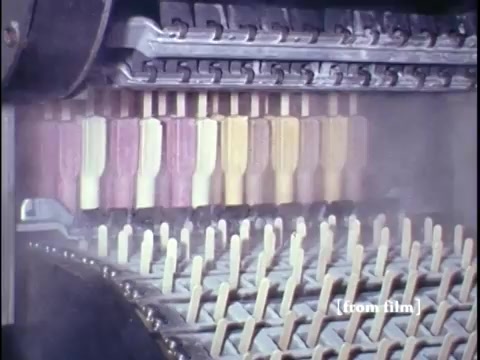 The still from 'Making Frozen Novelties - 1967 Kodachrome' showcases a detailed view of an industrial ice cream novelty production line. Visible are a series of popsicle molds, filled with colorful mixtures in various stages of preparation. The vibrant hues of the ice creams—ranging from creamy yellows to rich purples—are prominent. The machine appears to be automated, efficiently arranging the novelties on wooden sticks. The scene captures the essence of mid-20th century food production, highlighting the technology and processes involved in creating frozen treats.