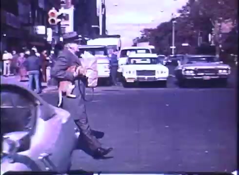 The still from the trailer of 'Harry and Tonto' features a man, presumably Harry, walking across a busy street while carrying a bag and holding a cat. The scene is set in an urban environment, with parked cars and pedestrians visible in the background, capturing a lively moment in the city. The colors are characteristic of a 16mm print, offering a nostalgic feel.
