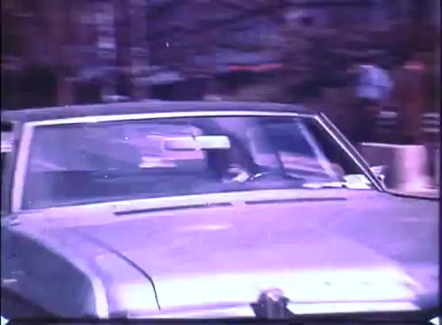 The still from the trailer of 'Harry and Tonto' features a vintage silver car driving through an urban setting. The scene captures a moment of movement, suggesting travel or adventure. The trailer likely emphasizes the themes of exploration and companionship in the film. The overall tone is nostalgic, fitting the 16mm print style.