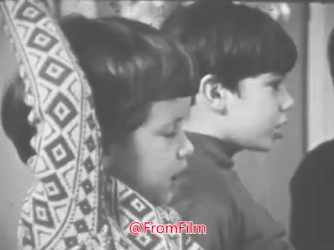 The still from the 16mm print of the 'Music Educators National Conference - 1969 PSA' features two children engaged in some sort of educational activity. One child appears to be wearing a patterned garment, while the other is closely observing or listening. The setting suggests a focus on music education, likely highlighting the importance of musical learning in youth development. The simplicity of the scene aligns with the period's style, conveying an earnest approach to educational themes.