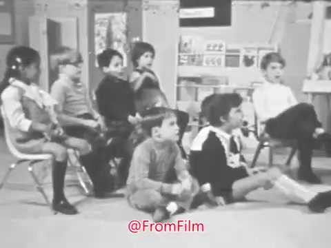The still from the 16mm print of the 1969 PSA for the Music Educators National Conference features a group of young children seated in a classroom setting. They appear engaged and attentive, indicative of a learning environment. The mix of boys and girls is dressed casually, reflecting the era. The backdrop includes educational materials, suggesting an emphasis on music education. Overall, the image captures a moment focused on children's involvement in music learning.