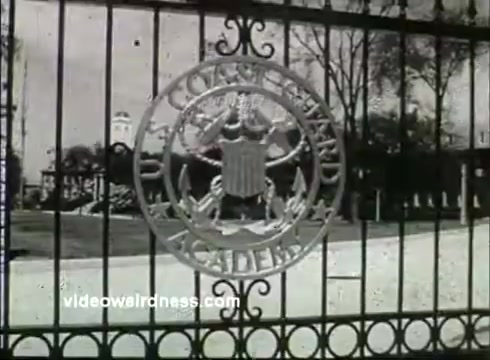 The still features a prominent metal gate with the inscription 'U.S. Coast Guard Academy' and the Academy's emblem. In the background, trees are visible, creating a serene campus atmosphere. The image evokes a sense of tradition and pride associated with the institution, characteristic of a 1950s public service announcement. The visual style reflects the era's film quality, typical of 16mm prints.