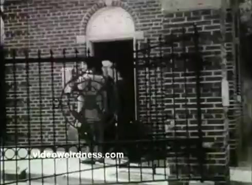 The still depicts a brick building entrance with a wrought iron gate featuring a life preserver symbol. Two individuals, likely military personnel, stand at the doorway. The scene captures a historical moment related to the Coast Guard Academy, typical of promotional material from the 1950s. The overall tone suggests a formal atmosphere, emphasizing the academy's nautical themes.