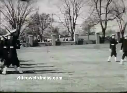 The still features a group of Coast Guard cadets in uniform, marching in formation on a grassy field. The background includes trees bare of leaves, typical of a 1950s setting, with onlookers visible near a brick boundary. The image has a classic black-and-white aesthetic, characteristic of 16mm film, conveying a sense of discipline and tradition associated with the Coast Guard Academy.