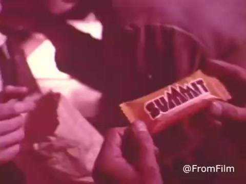 The still from 'Summit Cookie Bar - 1970's' features a person holding a brightly colored Summit candy bar wrapper, which is predominantly orange with bold, stylized text reading 'SUMMIT'. The background appears to be dimly lit, creating a nostalgic atmosphere typical of that era. The scene suggests a moment of sharing or displaying the candy, reflecting the product's appeal during its time of production in the early 1980s.