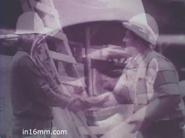 The still features two individuals, both wearing hard hats, shaking hands in a construction setting. One is standing on a ladder, while the other appears beside construction materials. The image has a vintage aesthetic typical of the 1960s, suggesting themes of teamwork and collaboration within the workplace. The context appears related to a public service announcement about safety or operational practices in construction.
