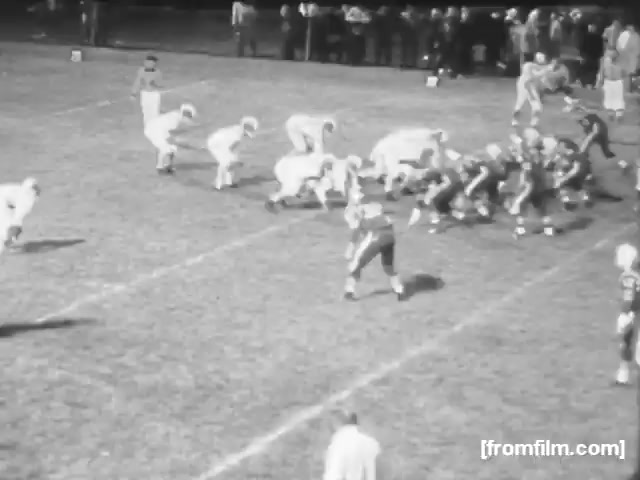 The still shows a high school football game in 1961, with players in traditional football uniforms. The field is marked clearly, and players are positioned for a play, indicating an active moment in the game. Spectators are visible in the background, suggesting an engaging atmosphere. The image captures the essence of high school sports during that era.