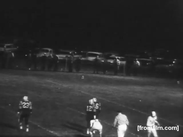 The still from 'High School Football - 1961' captures a night game scene. Two players, wearing jerseys numbered 12 and 23, are positioned on the field, while a referee in white observes the action. In the background, a crowd is lined up along the sidelines, and several vintage cars are parked, indicating a typical high school football atmosphere of the era. The black-and-white format adds a nostalgic feel to the image, reflecting the time period.