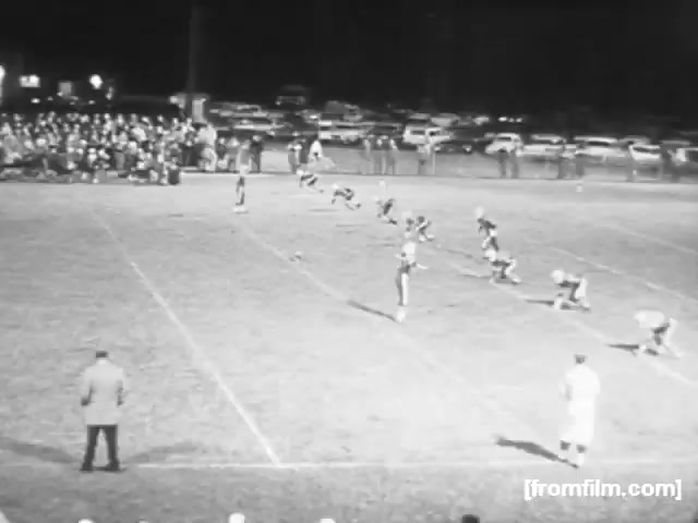 The still from 'High School Football - 1961' captures a nighttime football scene featuring players lined up for a kickoff. In the foreground, a coach or referee watches intently from the sideline. The field is lit, and a crowd is visible in the background, likely gathered in the bleachers. Cars are parked nearby, indicating a community event at a high school football game. The overall atmosphere reflects the excitement and tradition of high school football in that era.