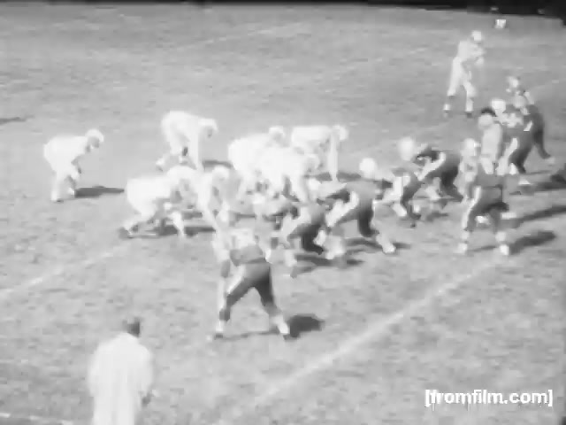 The still from 'High School Football - 1961' captures a moment on a football field, showing players lined up in formation. The scene presents a traditional high school football setting, with players wearing helmets and uniforms, engaged in the game. The field appears well-maintained, and the atmosphere suggests anticipation for the play to unfold. The film has a vintage quality, characteristic of the 16mm format.