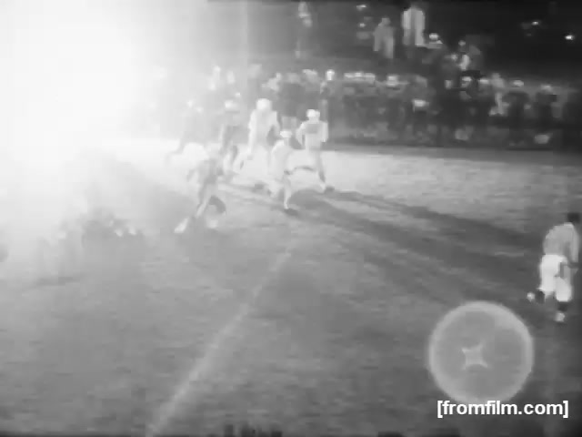The still shows a high school football game from 1961, captured in black and white on 16mm film. The field is illuminated with bright lights, suggesting an evening match. Players in helmets and uniforms are actively involved in a play, while the blurred outlines of spectators can be seen in the background. The overall atmosphere reflects the excitement and intensity of high school football during that era.
