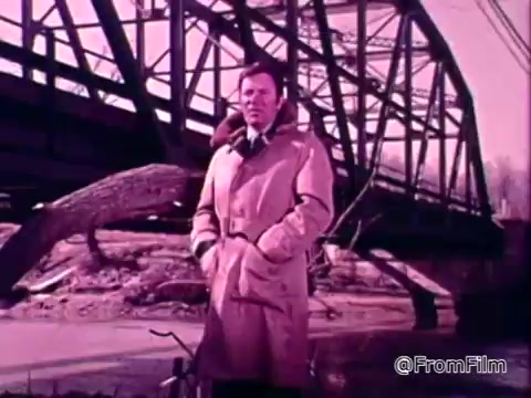 The still features a man standing by a river, likely after flooding. He wears a light-colored coat and appears serious, hinting at the gravity of the situation. In the background, a bridge looms, emphasizing the impact of natural disasters on infrastructure. The overall tone suggests a focus on raising awareness about the National Flood Insurance Program, which aims to protect communities from flood-related losses.