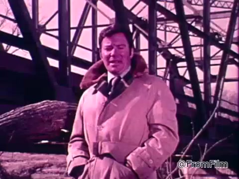 The still depicts a man, dressed in a tan coat with a fur collar, standing under a bridge or structure. The setting appears to be outdoors, likely after a flood, with debris visible in the foreground. The overall color tone is a washed-out pink, indicative of the film's vintage quality. This image is from a public service announcement featuring the National Flood Insurance Program and aims to inform viewers about flood protection options.